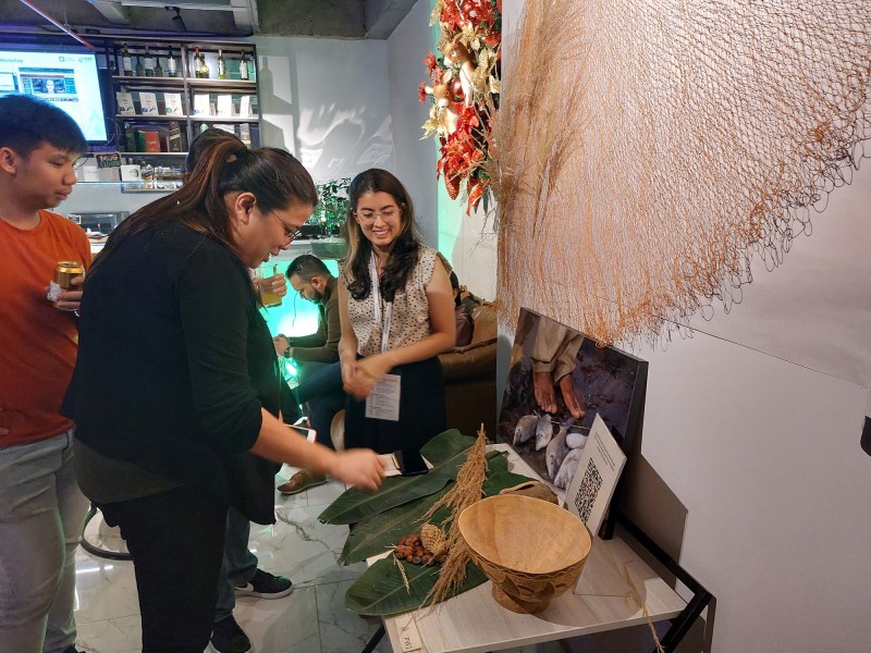 Three Hub members interactive with items in a display of props including large leaves and pieces of native flora, traditionally made bowls, a traditional fishing net, and an image of traditional fishing methods